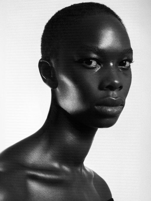 the image is a striking black and white portrait of a young woman with dark skin. she has a short, closecropped haircut and is shown from the shoulders up.  the lighting is dramatic, using strong contrasts of light and shadow (chiaroscuro) to highlight her features, particularly her intense gaze and defined bone structure.  her expression is serious and contemplative. the overall style is elegant and sophisticated, suggestive of high fashion photography.