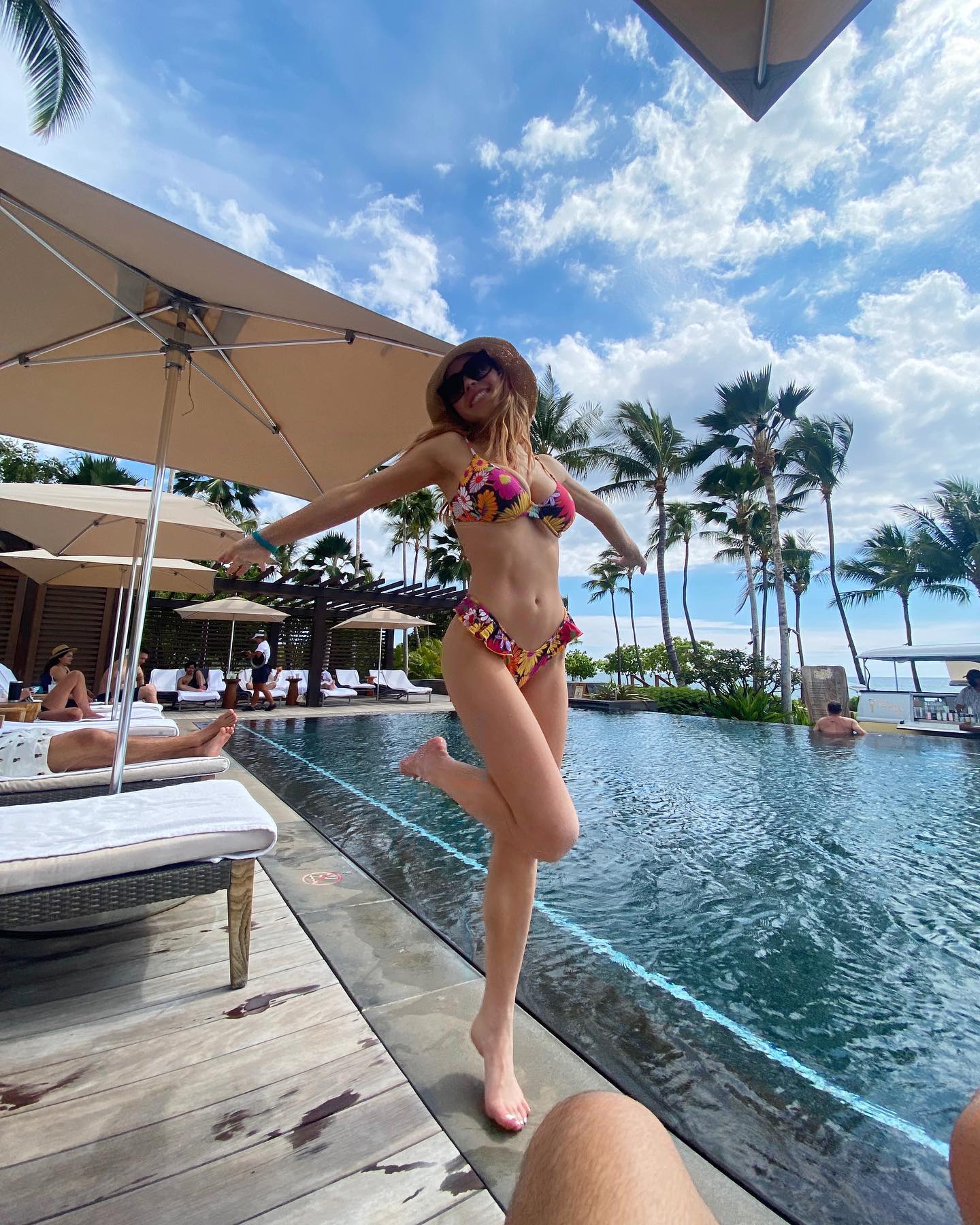 a woman in a colorful floral bikini smiles happily as she playfully stands by the edge of a resort pool.  the pool is surrounded by luxurious lounge chairs and palm trees, under a bright sunny sky. the scene is tropical and relaxing, suggestive of a luxury vacation.  a portion of another person's leg is visible in the bottom right corner of the frame.
