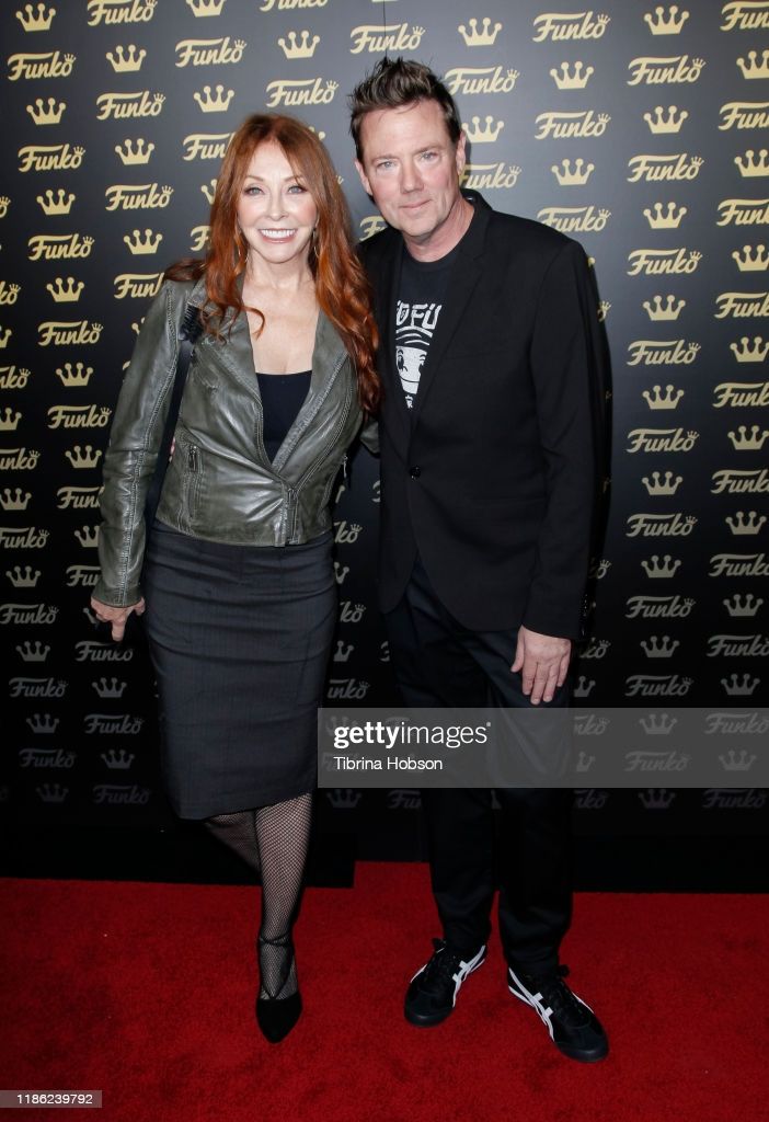 a man and woman pose on a red carpet for a premiere event. the man is wearing a suit and tie, while the woman is wearing a dress and jacket. they both have smiles on their faces. the background is a black and white wall with the word "funko" repeated many times.