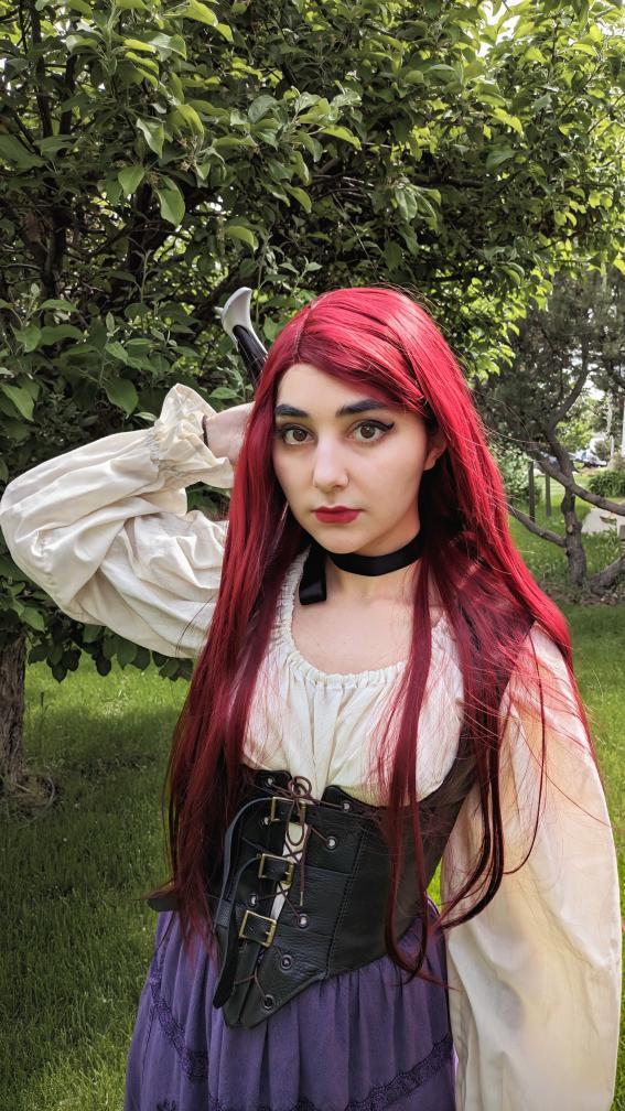 a young woman with long, vibrant red hair is posing outdoors in a cosplay outfit. she is wearing a creamcolored victorianstyle blouse, a black leather corset, and a purple skirt.  she has a darkcolored choker necklace and is holding a small dagger behind her head. the background is lush green grass and trees. the overall impression is a fantasy or medievalinspired costume.