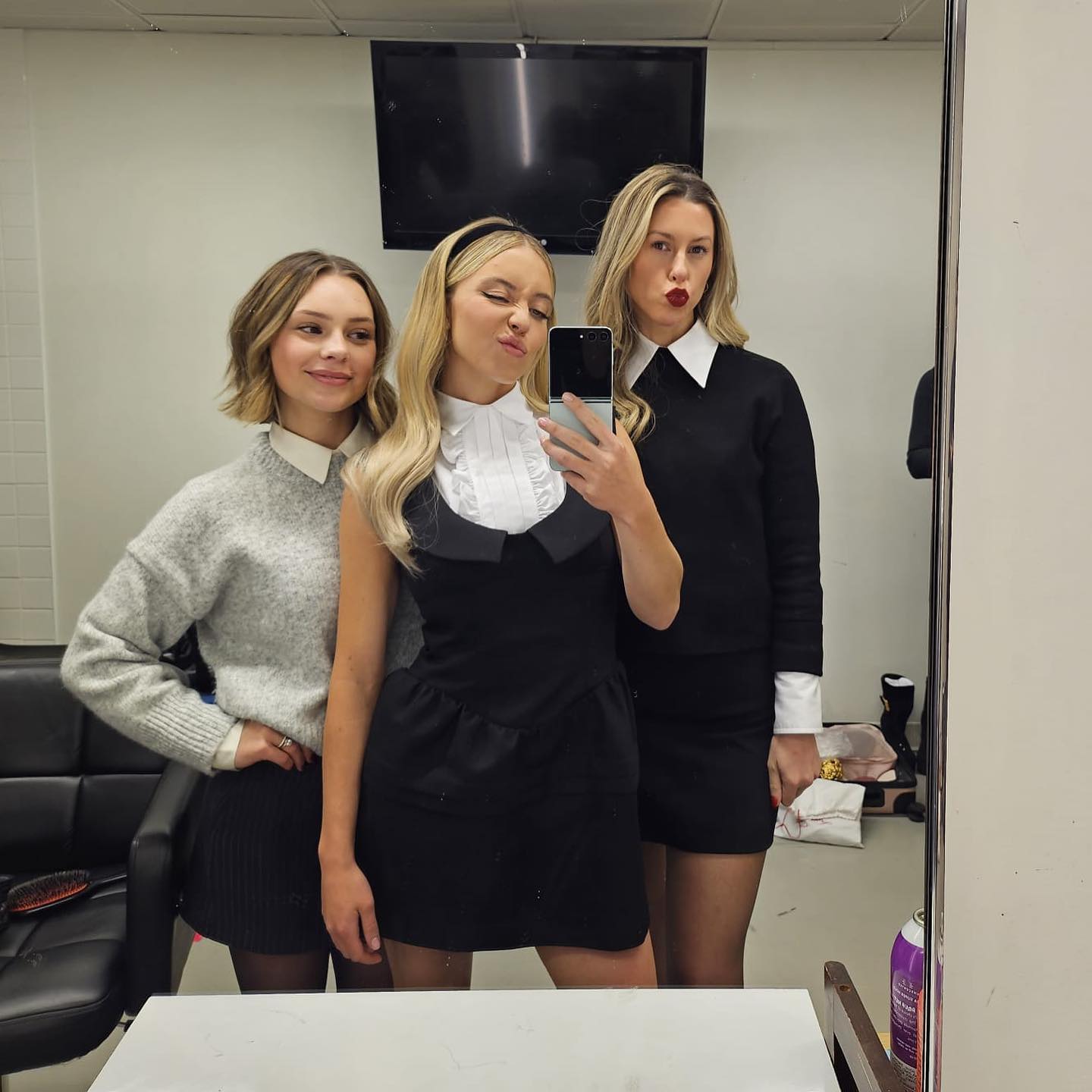 three young women, possibly actresses or television personalities, are taking a selfie in a dressing room mirror.  the woman in the center is wearing a black dress with a white peter pan collar and a headband. she's holding a phone to take the picture. on her left is a woman wearing a grey sweater and a black pleated skirt.  on her right is another woman in a black dress with a white collar. they appear to be backstage, possibly before or after a show or filming. the room shows typical backstage elements like a chair and a tv.  they're smiling and posing for the photo.