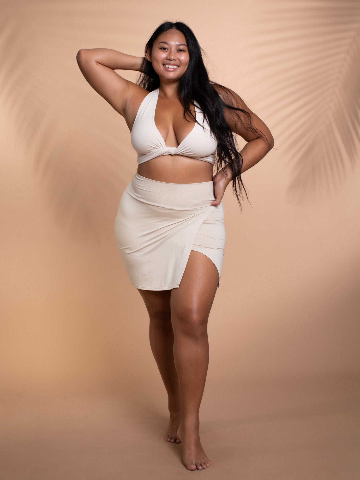 a fullbody shot of a confident, smiling plussize asian woman with long black hair. she is wearing a beige bikini top and matching wrap skirt. the setting appears to be a studio with a neutral beige background featuring palm leaf shadows. the overall mood is positive and celebratory of body diversity.