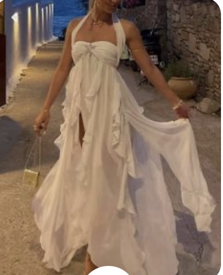 a woman in a white flowing halter dress is walking down a narrow cobblestone street. the dress has a highlow hemline and is adorned with ruffles. she is carrying a small gold purse and her hair is pulled back. the background suggests a seaside town, possibly in greece, with white buildings and a glimpse of the setting sun.