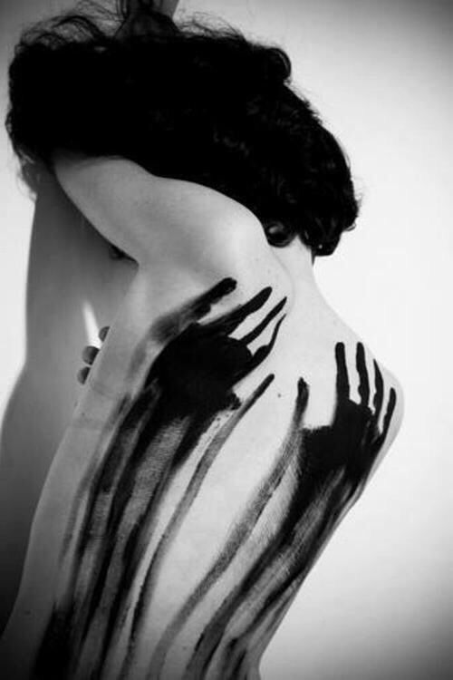 a black and white portrait of a woman with her back to the camera. her body is covered in black handprints, creating an abstract and mysterious design. the woman has long, dark hair that is partially covering her face. the image is shot in a studio setting, with a plain white background. the overall tone of the image is dark and dramatic.