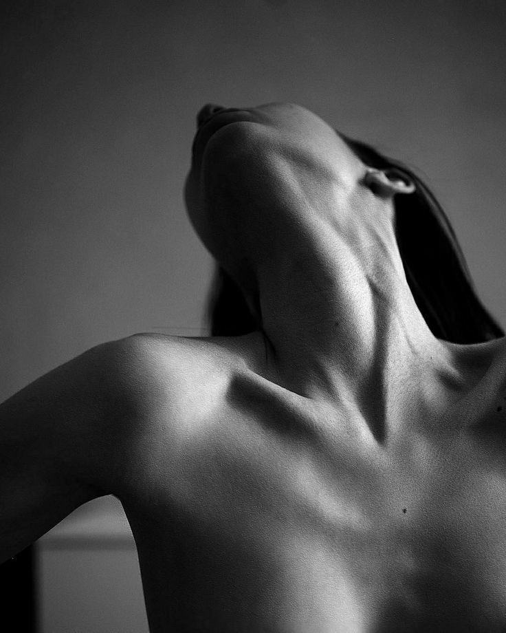 the image is a black and white closeup photograph of a woman's upper body, from the shoulders up.  she is nude, her head tilted back, arching her back slightly. the focus is on the delicate curves and shadows of her neck, shoulders, and collarbones. the lighting is dramatic, highlighting the texture and form of her skin. the overall mood is sensual and artistic, emphasizing the beauty of the human form. the simplicity of the black and white palette draws attention to the subject's physicality.