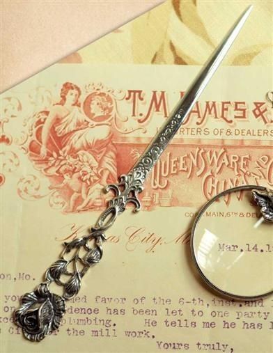 a silver colored letter opener in the shape of a rose laying on top of a vintage advertisement for t.m ames & co, importers and dealers of queensware and china. the letter opener is shaped like a stem of a rose with leaves and a bloom at the bottom. the top is a smooth, pointed blade. there is also an antique letter with a portion of the writing visible.