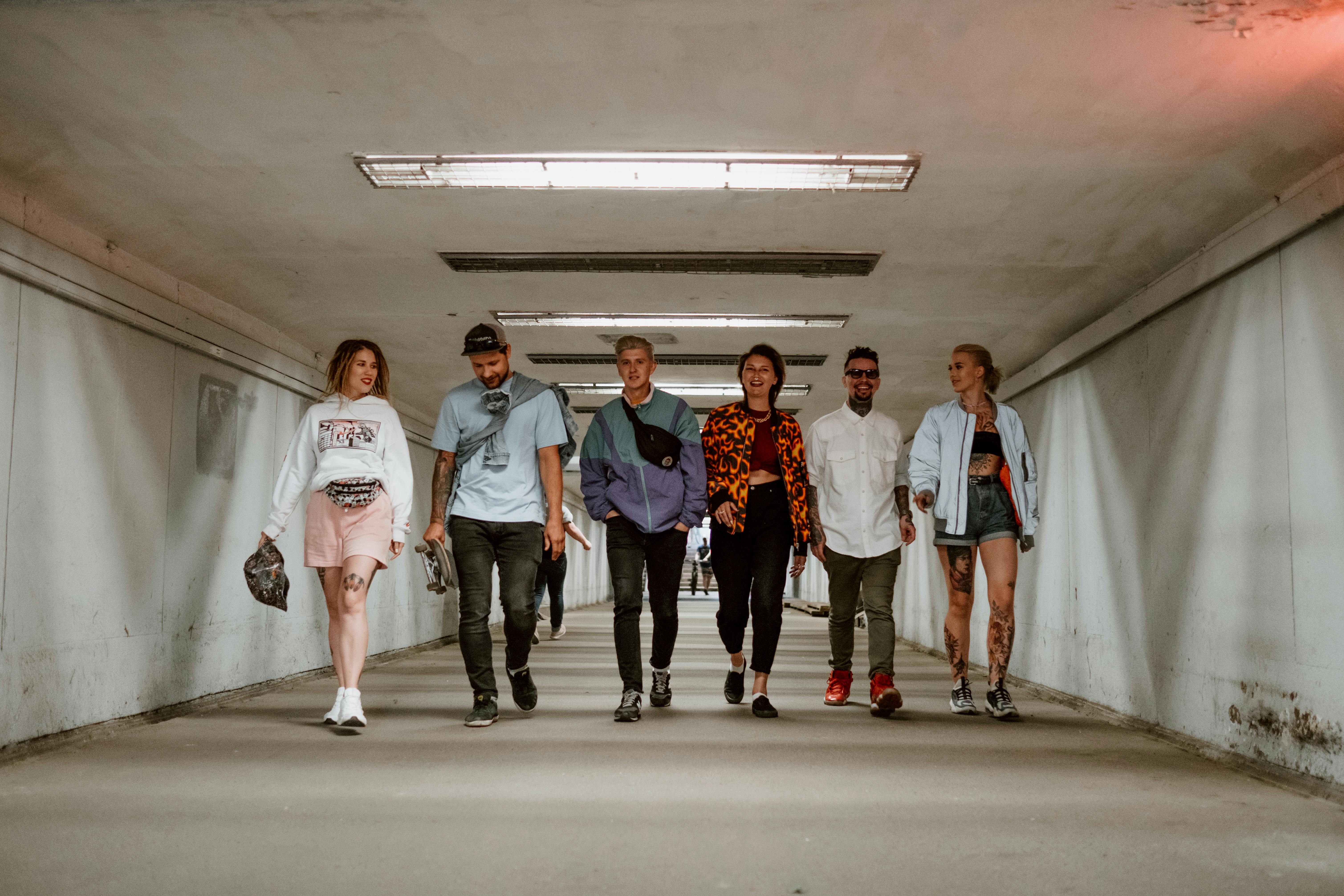 the image shows a diverse group of seven young adults, four men and three women, casually dressed and walking down a long, brightly lit concrete underpass or tunnel.  the group is positioned centrally in the frame, walking towards the viewer. the individuals exhibit a range of styles, with some showcasing tattoos and various types of casual clothing, including jackets, tshirts, shorts, and pants. one person is carrying a skateboard. the overall ambiance is relaxed and friendly, portraying a sense of youthful camaraderie. the setting is urban and modern.