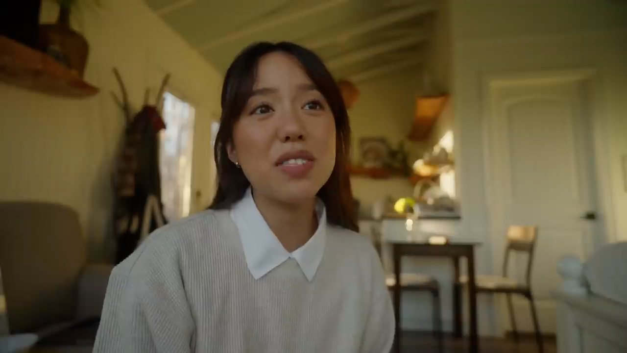 a young asian woman sits in the living room of her cozy and minimalist tiny home. the warm, natural light coming through the windows illuminates the space, creating a peaceful and inviting atmosphere. the woman is looking offcamera with a thoughtful expression on her face. the color palette is dominated by warm, earthy tones, further enhancing the cozy and inviting feel of the scene.