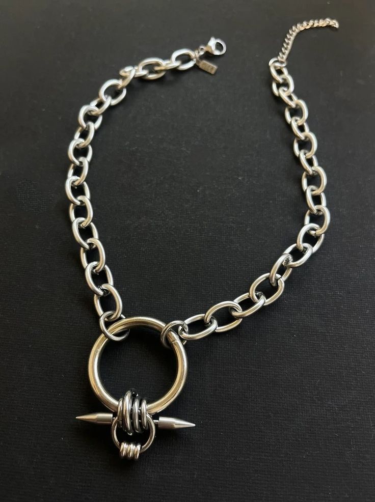 a silver chain necklace with a circular pendant that has two spikes sticking out. the chain is made of large, thick links and the pendant has a slightly distressed finish. the necklace is laying flat on a dark background.