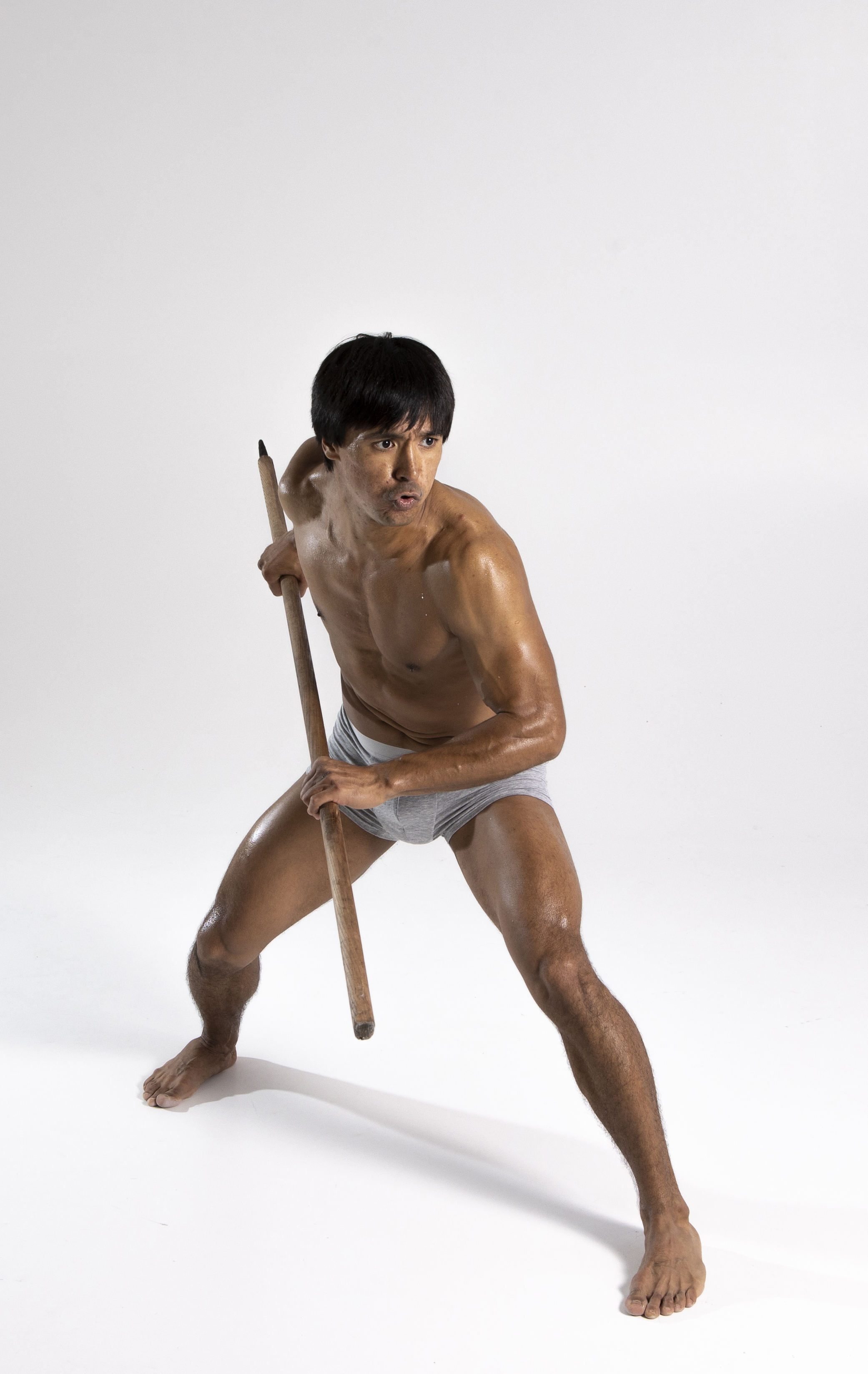 a barechested man in gray underwear is holding a wooden stick in both hands. he is barefoot and standing in a defensive stance with his legs spread apart. he is looking away from the camera and appears to be indoors against a white background. he has dark hair and a muscular build.