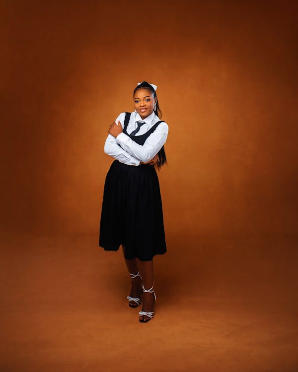 a fullbody studio shot of a woman with her arms crossed, wearing a white blouse, a black skirt, a tie, heels, and a headband, standing against a brown background.
