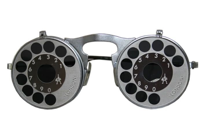 the image shows a pair of vintage eyeglasses with rotary lenses. each lens housing is circular with numbered positions around a central viewing area. the numbers range from 0 to 9, with a small symbol and the word "london" on the lower part of each housing. the glasses are silvercolored, with a bridge connecting the two lens housings. the lenses appear to be adjustable with different black inserts that can be rotated into place. they are used for eye examinations, specifically for measuring refraction and determining a patient's prescription.