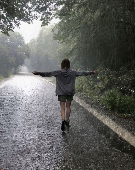 a young woman walks away from the camera down a rainsoaked road that winds through a lush, green forest.  the lighting is dim and overcast, creating a moody and atmospheric scene. the woman's arms are outstretched as if embracing the rain.