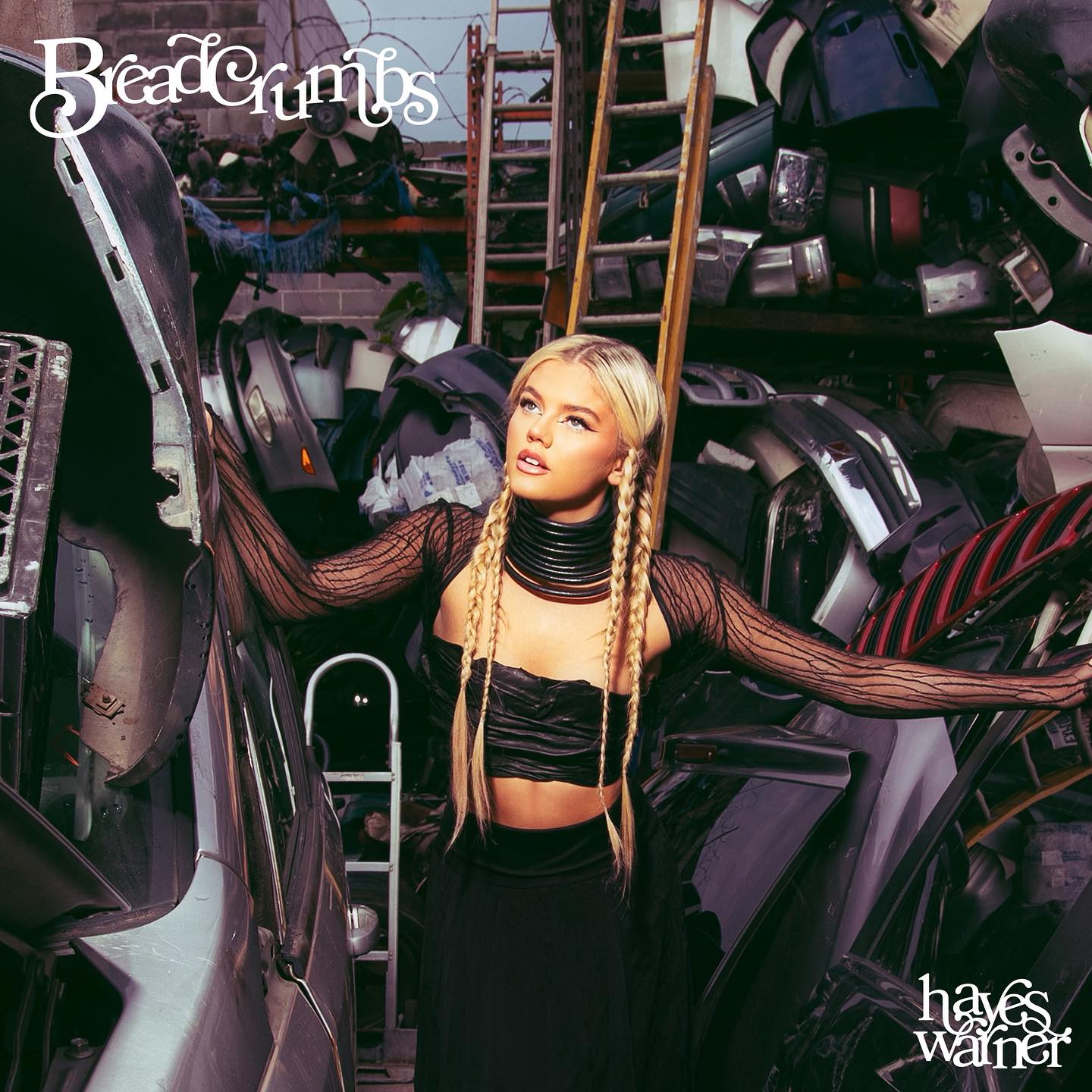 the image is the album cover for hayes warner's "breadcrumbs." a young woman with long blonde hair styled in braids is the focal point, wearing a black crop top and long skirt, and a thick black choker. she is positioned among car parts in what appears to be a junkyard, with various metal and automotive components stacked around her. a ladder is visible in the background. the album title "breadcrumbs" is written in a stylized font in the top left corner, and the artist's name "hayes warner" is in the bottom right corner.