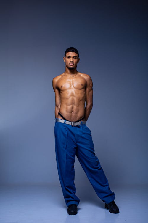 a portrait of a muscular man wearing only blue pants, a black thong, and black shoes in a studio setting. he has tan skin and is wearing a gold chain around his neck. he is posing with his hands behind his back.