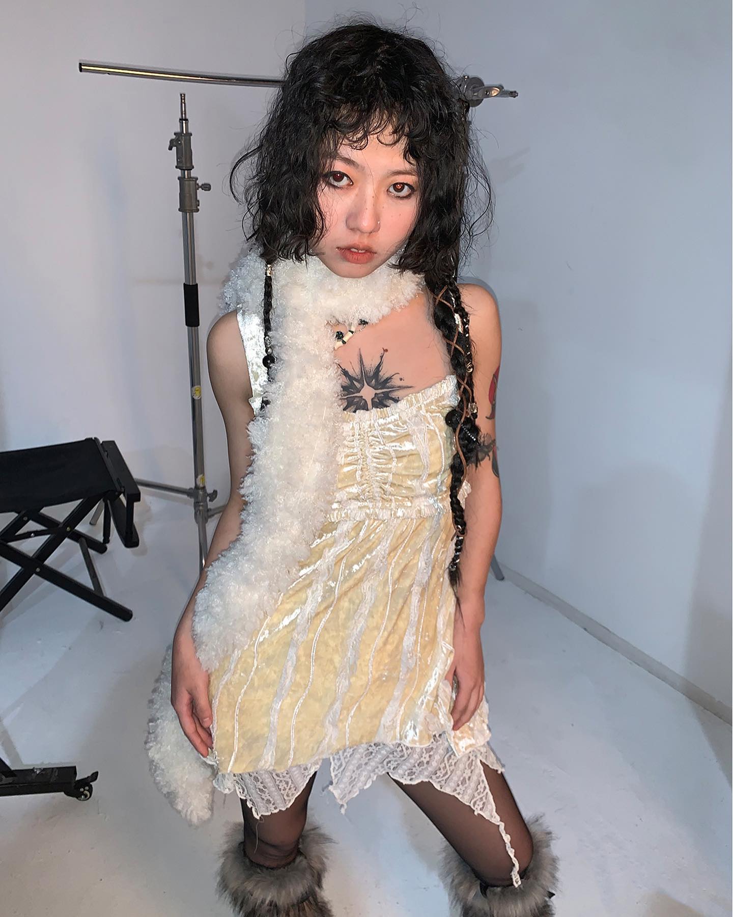 a young woman with short, dark curly hair and bangs poses in a studio setting. she's wearing a pastel yellow velour dress with lace trim at the hem. a white faux fur stole is draped around her neck and shoulders. she's also wearing black tights, with a hint of fishnet visible. the woman has a star tattoo on her chest. the background is a plain white wall. a photography setup with lighting equipment is partially visible in the background.  her style leans towards alternative, grunge, or indie fashion.