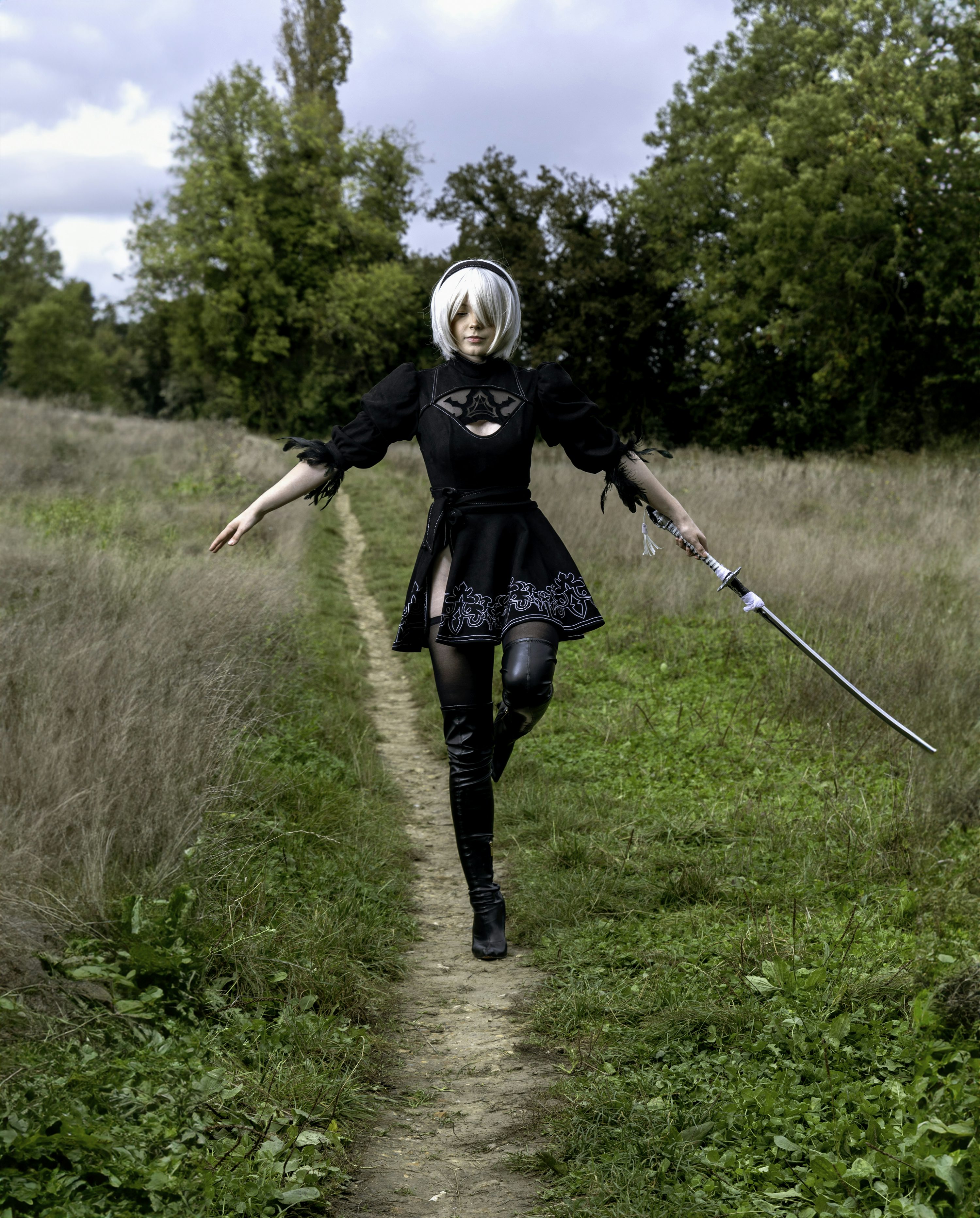 a woman cosplaying as 2b from nier: automata is walking along a path in a field. she is wearing a black dress, thighhigh boots, and a white wig with a headband. she is holding a sword in her right hand. the path is surrounded by tall grass and trees. the sky is cloudy.