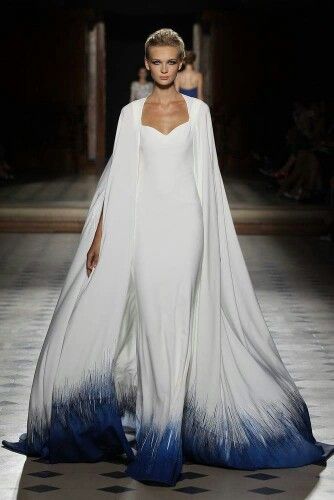 a fashion model walks the runway in a long white gown with a dramatic cape. the dress is trimmed with a blue ombre pattern at the bottom, which adds a touch of color and whimsy to the otherwise simple design. the model has her hair pulled back, and she is wearing minimal makeup, allowing the dress to be the center of attention. the runway is lined with spectators, eager to see the latest designs.