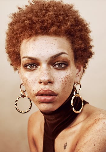 a closeup portrait of a woman with freckles, short brown afro, and brown eyes. she is wearing large, gold, circular earrings and a brown turtleneck sweater. a small chest tattoo is visible.