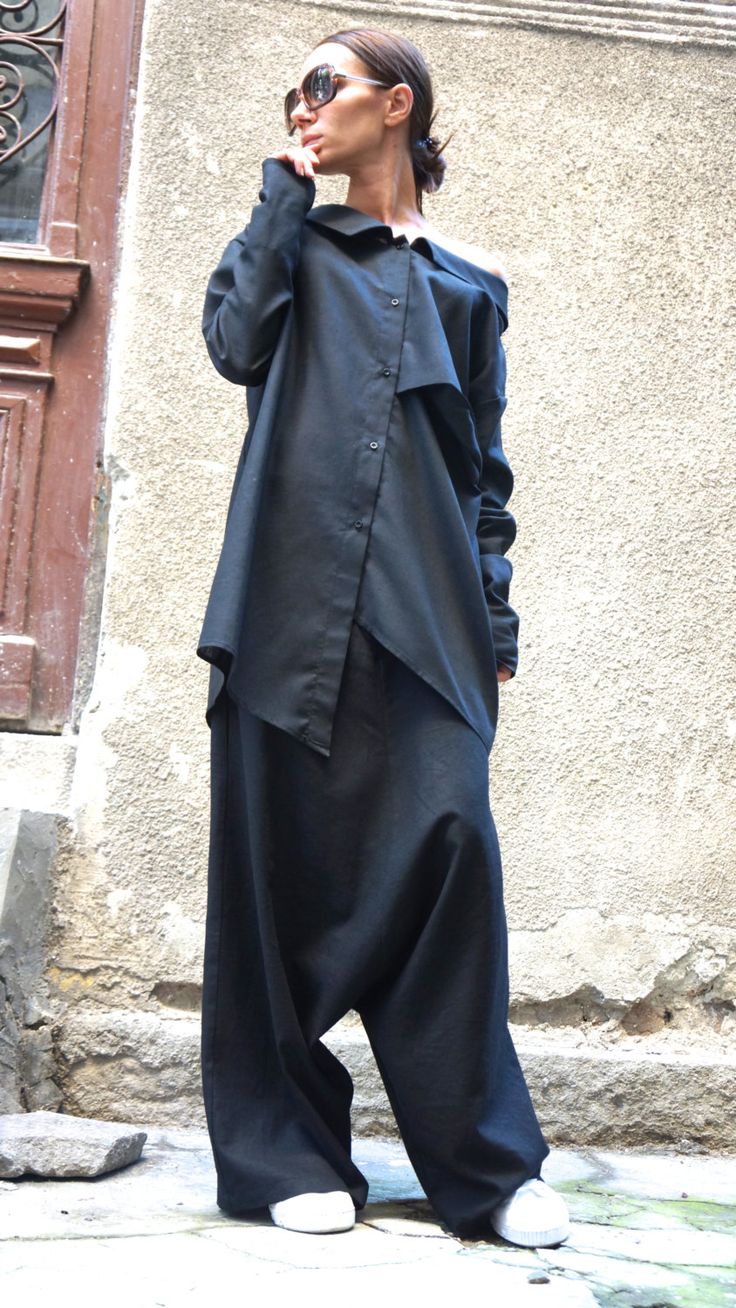 a woman stands outside wearing a black, offtheshoulder button up shirt and baggy black pants with white sneakers. she has sunglasses on and is turned to the side, looking off into the distance.