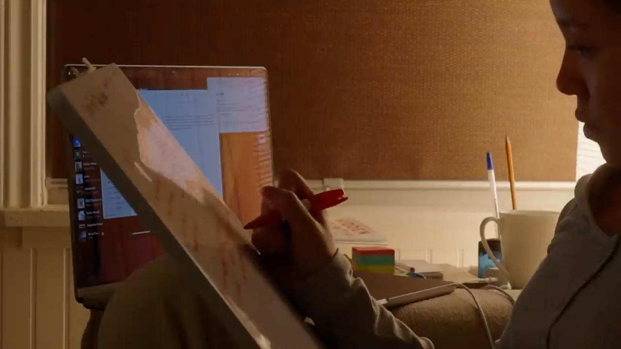 a woman is sitting at a desk lit by warm lamplight, working on a drawing pad propped up against her laptop.  the perspective is from over her shoulder, giving a sense of her focus and concentration on the task. there are signs of work and study around her, suggesting a home office or study space.