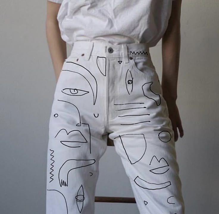 a woman is wearing custom white highwaisted jeans with black line art drawings of abstract faces and shapes. she is also wearing a white tshirt. she is sitting on a stool. the background is a plain, lightcolored wall.