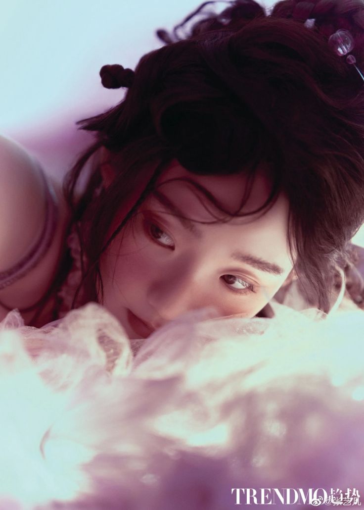 this is a photo of a woman with long black hair, wearing a white dress and  jewelry,  posing in a studio. she is looking away from the camera with a soft, dreamy expression. the lighting is soft and creates a romantic atmosphere.  the image is in focus on the woman's face and hair.  she is surrounded by a white backdrop and soft light. the image appears to be an editorial shoot for a magazine.