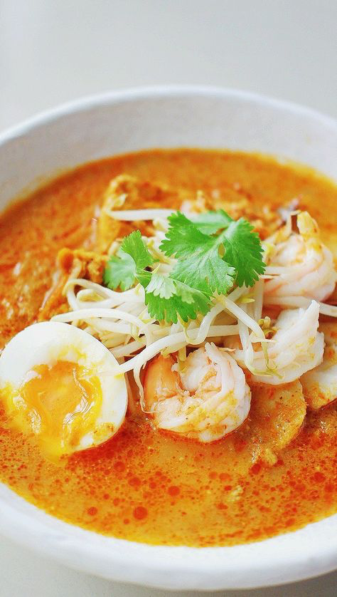 a closeup shot of a bowl of laksa, a popular southeast asian noodle soup. the laksa is a rich, orangered curry broth filled with shrimp, a softboiled egg, bean sprouts, and garnished with fresh cilantro. the bowl is white, and the background is a blurred, lightcolored surface. the image is welllit and appetizing.