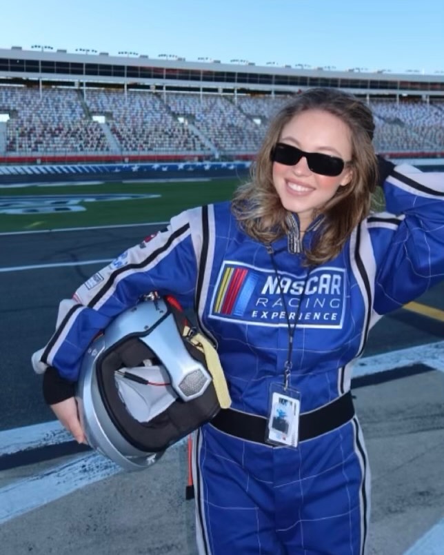 a young woman, wearing a blue nascar racing experience jumpsuit and sunglasses, holds a racing helmet while standing at a race track.  the grandstands of the speedway are visible in the background.  the image suggests a fun, exciting atmosphere.