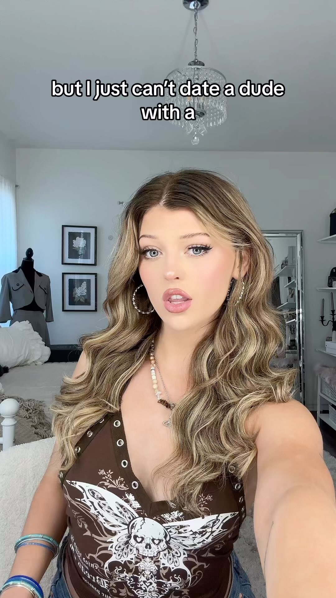 a young woman with long curly blonde hair is centered in the frame. she is wearing a brown top with a skull design, a necklace and hoop earrings. behind her are various objects, including a bed, a chandelier, pictures on the wall, and a clothing rack with a tailored outfit. the text "but i just can't date a dude with a" is overlaid on the image.
