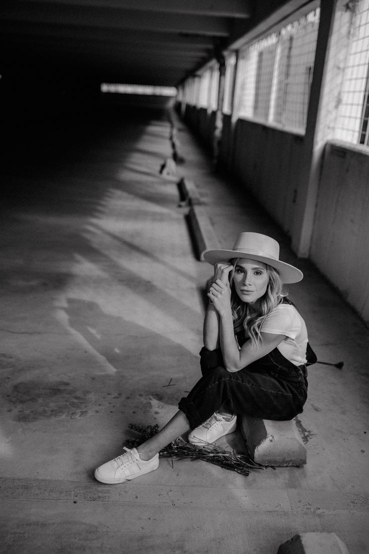 a black and white photo of a woman sitting on a concrete block in a dimly lit alleyway. she is wearing a widebrimmed white hat, denim overalls, and white sneakers.  she has her hands near her face, appearing pensive or thoughtful. the setting is industrial and gritty, with shadows adding to the moody atmosphere. the overall aesthetic is vintage or retro.