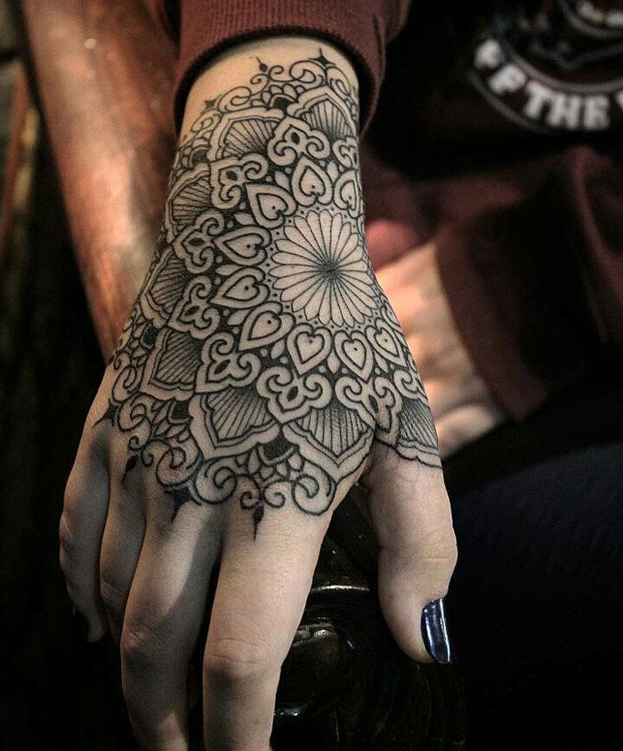 a closeup shot of a person's hand and wrist adorned with an intricate black ink mandala tattoo. the tattoo covers the back of the hand and extends onto the wrist. the design features a flowerlike element in the center, surrounded by various patterns and shapes including heartlike motifs. the person has a dark blue nail polish on their thumb. the overall style resembles a henna tattoo.