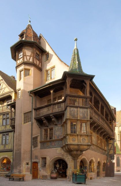 a view of a historical building in colmar, france. the building is made of stone and wood, with a distinctive curved facade and a tower. the building has multiple levels, with balconies and windows on each level. the street in front of the building is paved with stone. the sky is blue and clear.