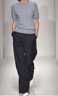 a woman is wearing a light grey, longsleeved tshirt, paired with black, widelegged pants. she is wearing white sneakers and is posing on a runway.  the photo appears to be taken from a fashion show.