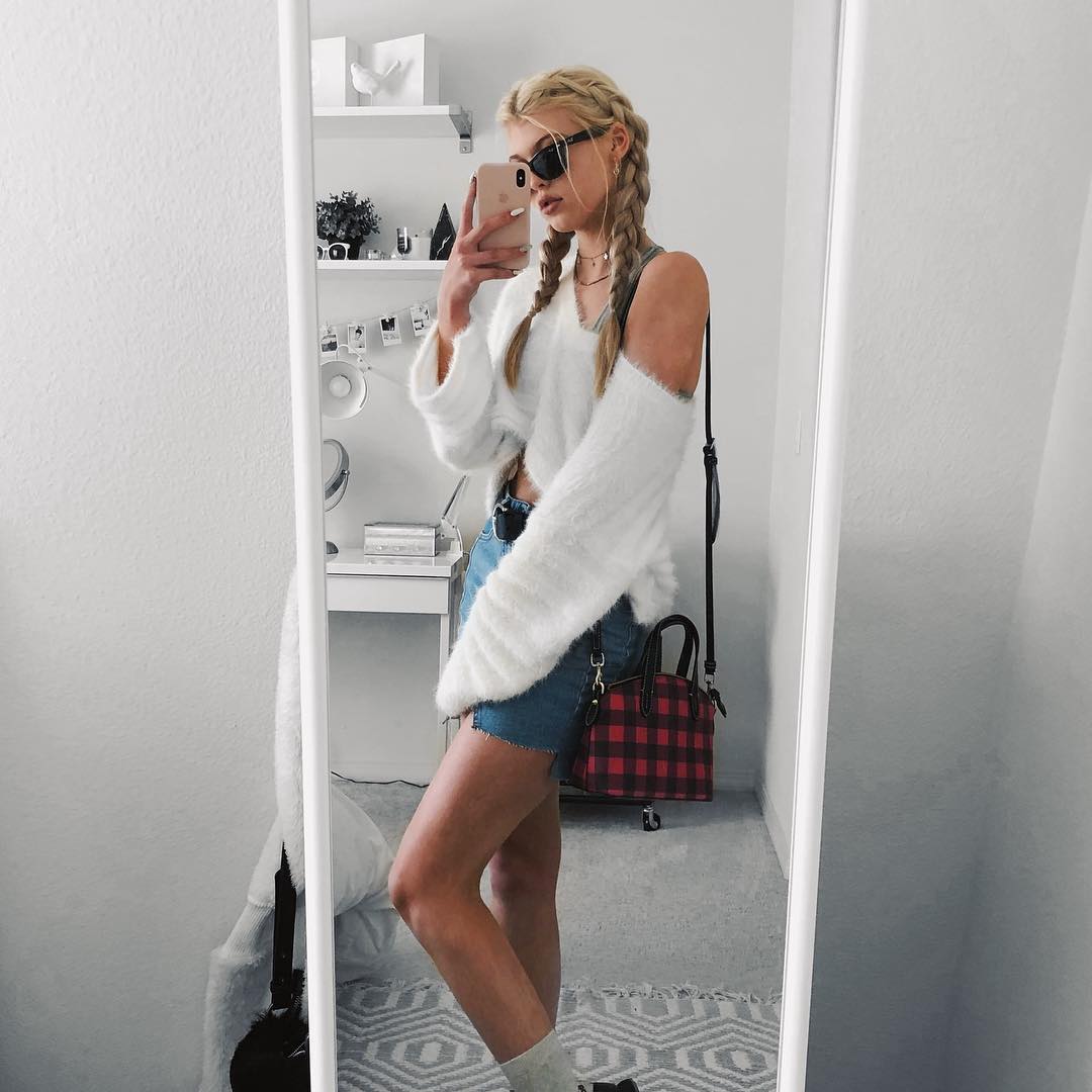 a woman with blonde hair styled in braids stands in front of a mirror, taking a selfie with her phone. she is wearing sunglasses, a white sweater with a cold shoulder design, and denim shorts. a red plaid purse hangs from her shoulder. the reflection shows a shelf with decorations, and the room is brightly lit. a rug is visible on the floor.