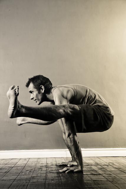 a black and white portrait of a man doing a handstand with his legs extended in front of him. he is in the middle of a yoga pose, and appears to be in good shape. the background is a plain wall, and the floor is wooden.