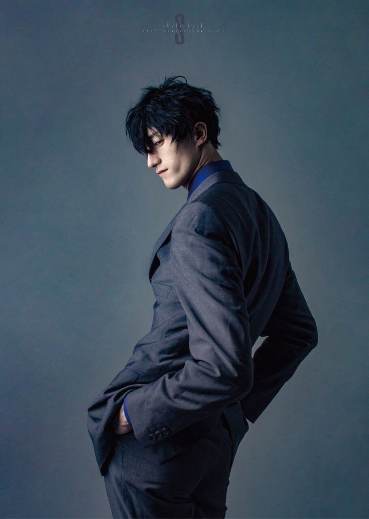 a man in a grey suit is standing against a grey background. he is wearing a blue shirt and a tie. he has his hand in his pocket and his head is tilted down. the man has short black hair and is looking thoughtful. he is posed in a professional and stylish manner.