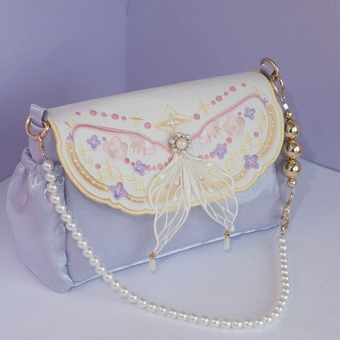a white and purple handbag with an embroidered butterfly design and a pearl strap. the butterfly has pink and purple details, and there are small pearls hanging from its wings. the bag is made of velvet and has a chain strap with pearl accents.