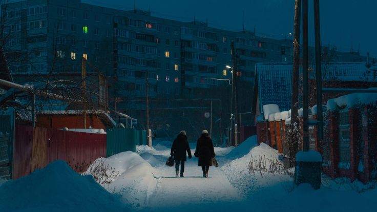 the image depicts two individuals walking down a snowcovered street at night in a residential area. the street is lined with snowcovered fences, and the buildings in the background are dark apartment buildings with a few lights visible in the windows. the scene is cold, dark, and quiet, with a serene winter atmosphere. the people appear bundled in winter clothing, walking together and carrying bags.  the overall tone is somber and peaceful.