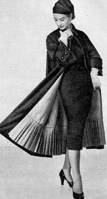 a monochrome image shows a woman in vintage fashion. she is wearing a fitted dress and a coat with a pleated hem. she also has a hat, gloves, high heels, and jewelry.