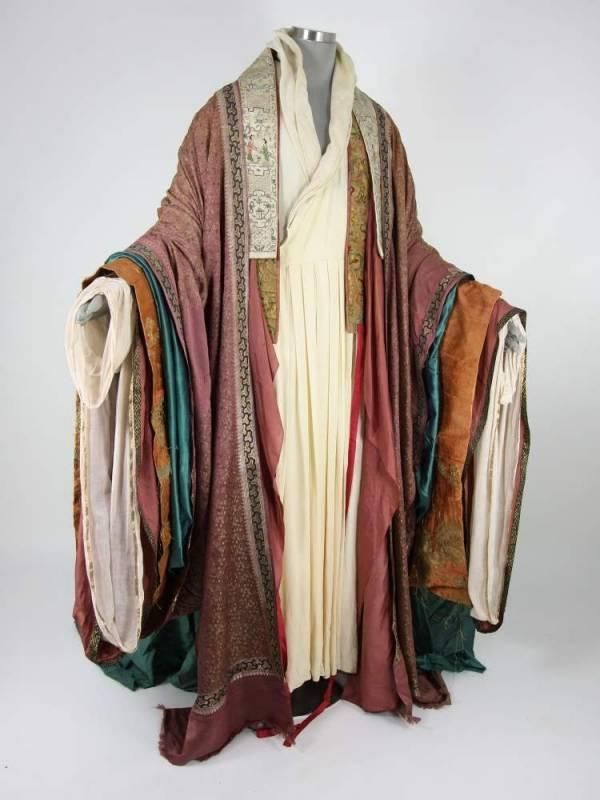 a photograph depicts a mannequin dressed in an elaborate multilayered robe against a plain white backdrop. the robe consists of a variety of colors, including maroon, green, orange, and ivory, creating a rich and textured appearance. the maroon fabric appears to form the outer layer, while the green and orange elements add depth and contrast. the ivorycolored fabric is more prominent at the front, suggesting an underlayer or inner garment. intricate trim detailing can be observed on several layers, lending an oriental or formal aspect to the attire. a decorative collar further enhances the outfit's formal nature, making it a visually striking and intricately designed piece of clothing.