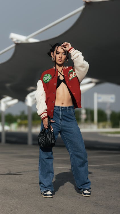 a young woman stands confidently in a parking lot, posing for a fashion photo. she wears a red and white varsity jacket, loosefitting blue jeans, and black and white saddle shoes.  she carries a small black purse and has her hair styled in braids. the background features a modern architectural structure with a large fabric canopy. the overall style is urban and trendy.