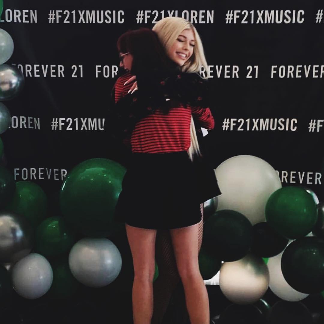 two women are embracing in front of a backdrop with "forever 21" branding and hashtags, surrounded by balloons. one woman has blonde hair and is smiling, while the other has reddishbrown hair and is wearing glasses. the backdrop is black with white text, and the balloons are in shades of green, silver, and white. the woman with blonde hair is wearing a red and black striped shirt and a black skirt, while the other woman's outfit is partially obscured.
