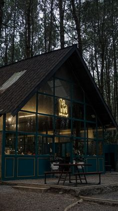 an image of a cabinstyle restaurant nestled in a forest at night. the cabin is illuminated from within, showing large glass windows that showcase the interior. there are tables and chairs set up outside on a gravel patio. a neon sign with a logo is visible on the glass.  the overall atmosphere is dark and moody, with the lights of the restaurant contrasting against the dark trees.
