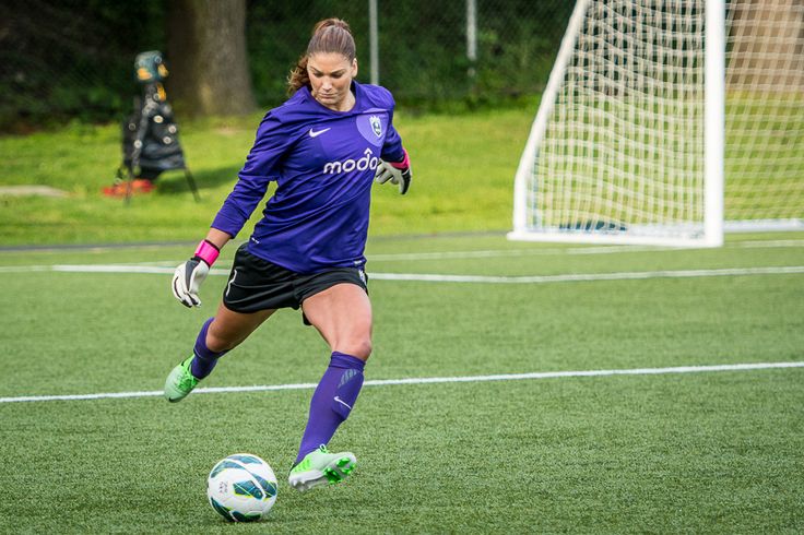 a female soccer goalkeeper in a purple uniform makes a dramatic save, her body extended as she kicks the ball away from the goal. the image captures the intensity and athleticism of the sport. the bright, sunny day illuminates the green soccer field.