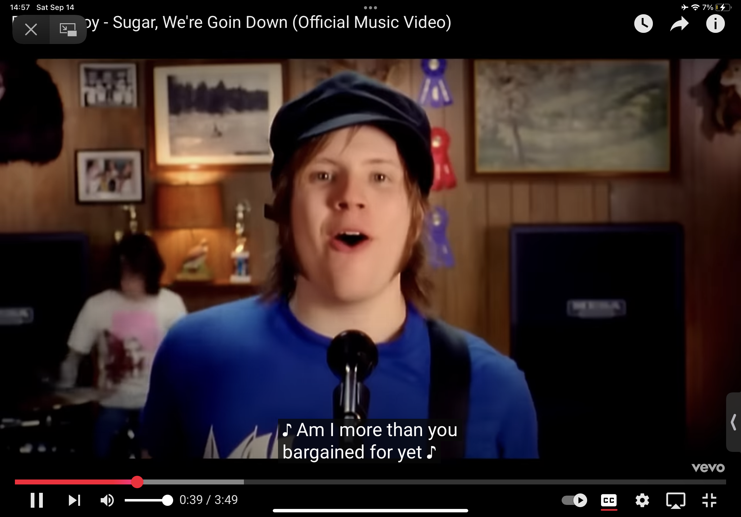 a screenshot from a music video featuring a young man with a microphone singing. he appears to be on some type of stage, likely in a basement setting, surrounded by awards and musical equipment. the lighting is bright, suggesting stage lighting. the overall aesthetic hints at a pop punk or emo genre, popular in the 2000s. the text overlay reads "am i more than you bargained for yet" which are lyrics from the song.