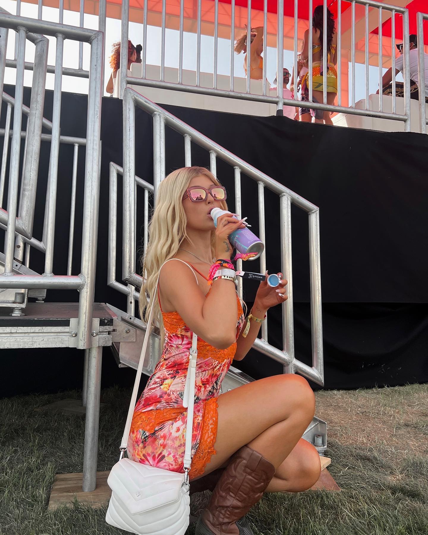a woman in a colorful floral dress, sunglasses, and brown boots is squatting and drinking from a water bottle. she is wearing several bracelets and has a white purse slung over her shoulder. she is positioned in front of a metal staircase and structure. in the background, people can be seen on a platform and in a crowd, suggesting she is at an outdoor festival.