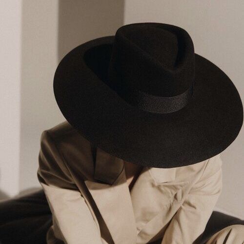 a woman is wearing a wide brim black fedora hat and a beige blazer. the image is cropped from the shoulders up. the light background is mostly out of focus and the focus is on the woman and her hat.