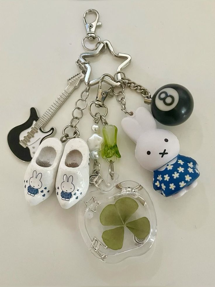 a keychain with various charms attached. the charms include a miffy bunny figurine, an eight ball, a guitar, a pair of wooden shoes/clogs, and a clover encased in resin. the keychain itself is made of silver and shaped like a star.