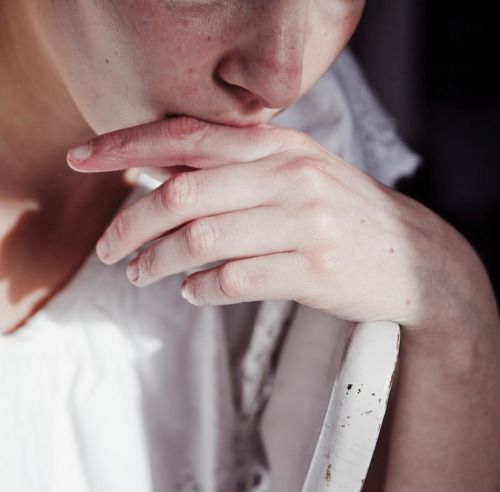 a closeup portrait of a woman with fair skin and freckles. she is wearing a white dress and her hand is raised to her mouth, partially obscuring her lips. the lighting is soft and natural, highlighting the texture of her skin and the delicate fabric of her dress. the overall feeling of the image is one of intimacy, vulnerability, and contemplation.